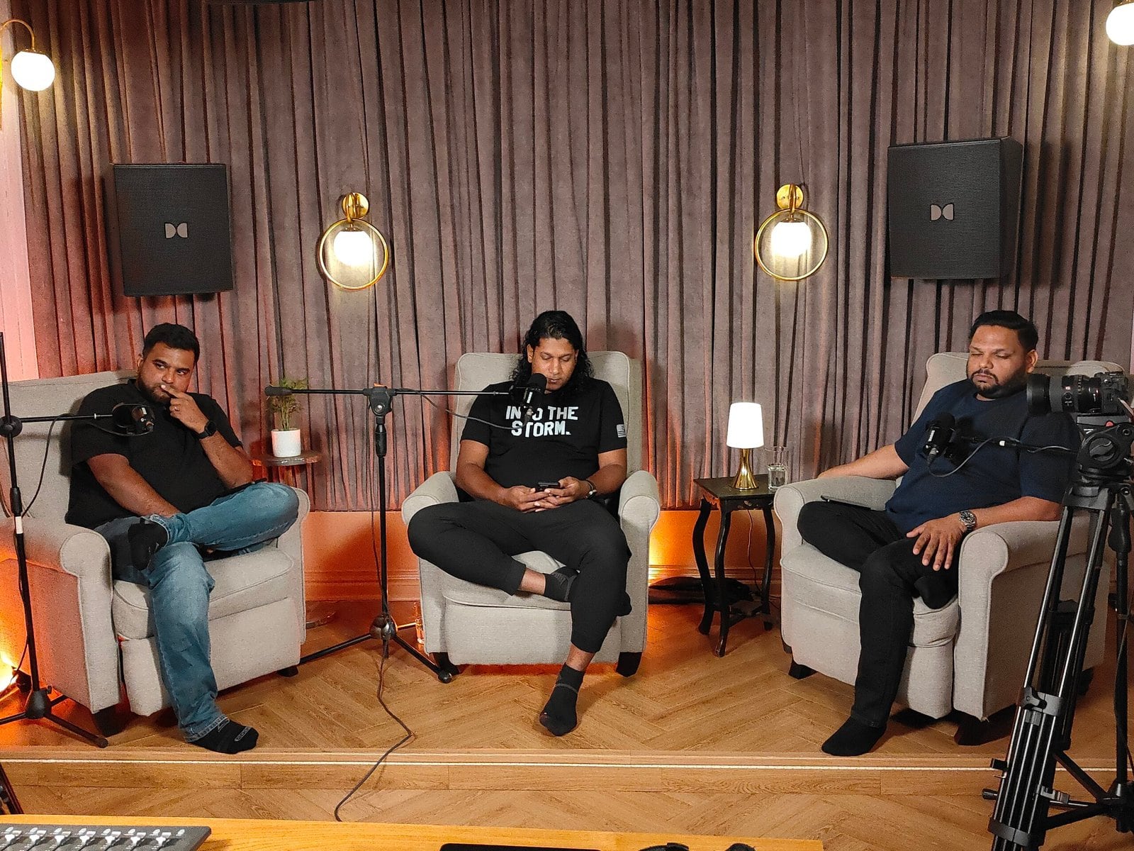 A three-person panel recording on the podcast set, with boom microphones and a camera in the foreground