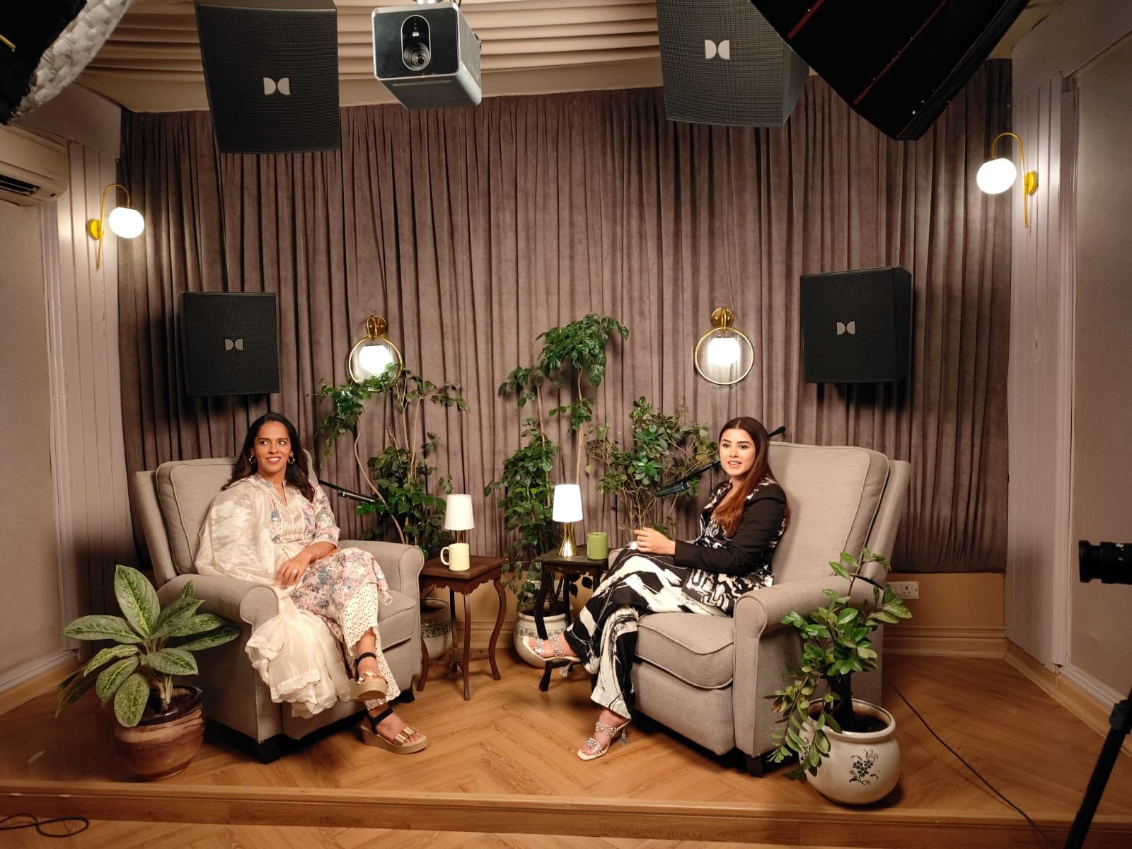 Two guests in conversation on the podcast set among potted plants, lit by a pair of wall sconces